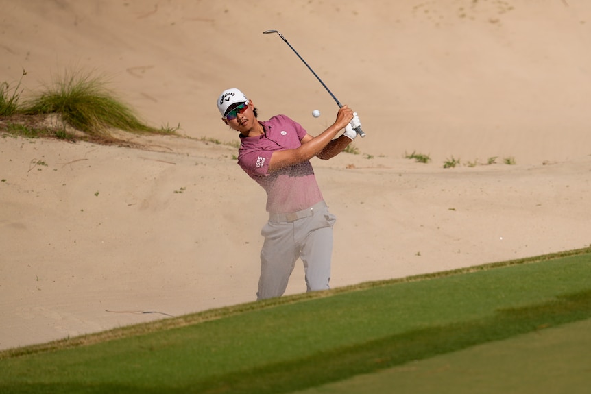 Min Woo Lee plays a shot out of a bunker, with the ball seen between his face and the golf club