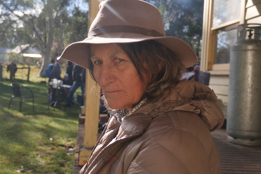 A woman in a hat and coat leans on a verandah and looks at the camera with a serious expression