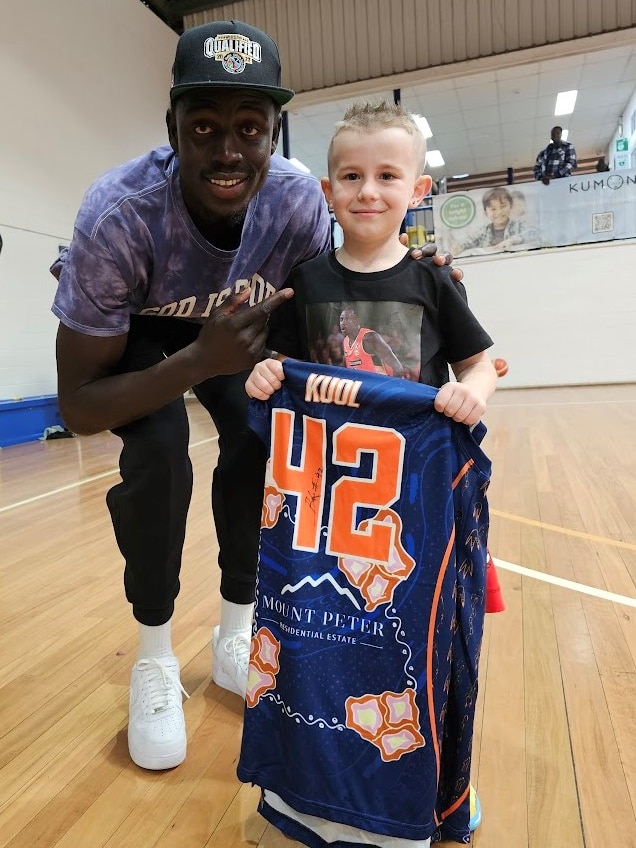 A man wearing a cap nad blue tee bends down beside a young buy holding a basketball singlet. 