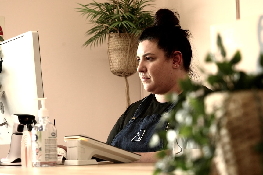 A woman in a black T-shirt, her dark hair swept up into a bun, sits in front of a computer at a counter