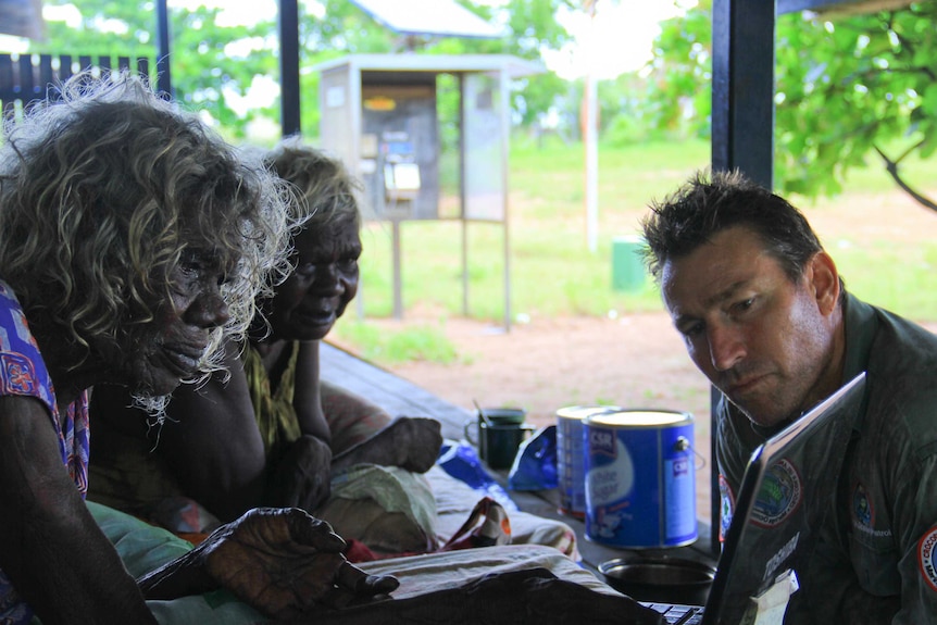 Indigenous mal and female sitting with park ranger in front of laptop, greenery in background.