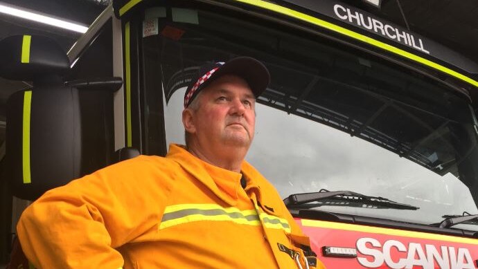 A firefighter volunteer wearing bright yellow and a cap stands beside a fire truck