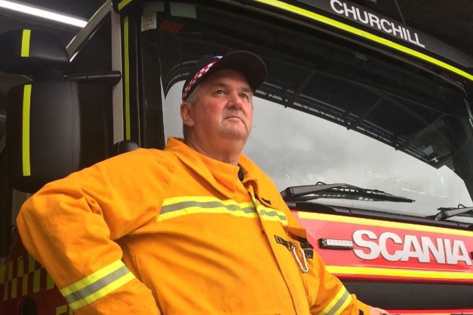 A firefighter volunteer wearing bright yellow and a cap stands beside a fire truck