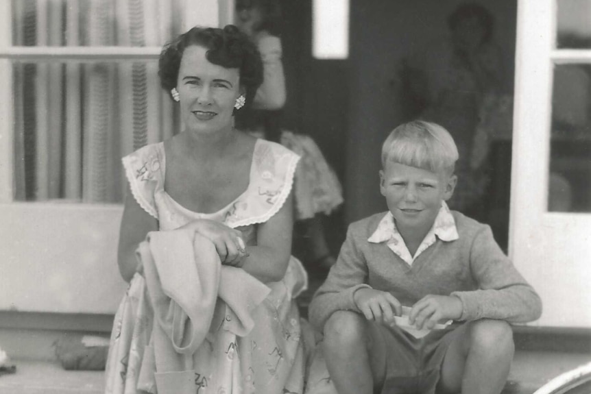 A black-and-white photo of a mother and her child sitting on the front porch of a house, circa 1960s.