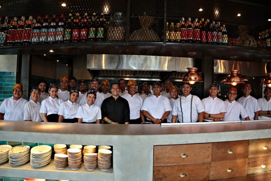 A man in black is surrounded by people in white with a huge stainless steal kitchen behind them.