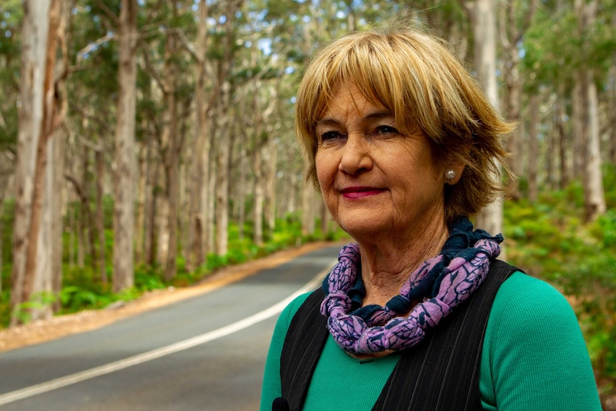 A woman stands in front of a road. She is surrounded by trees and greenery.