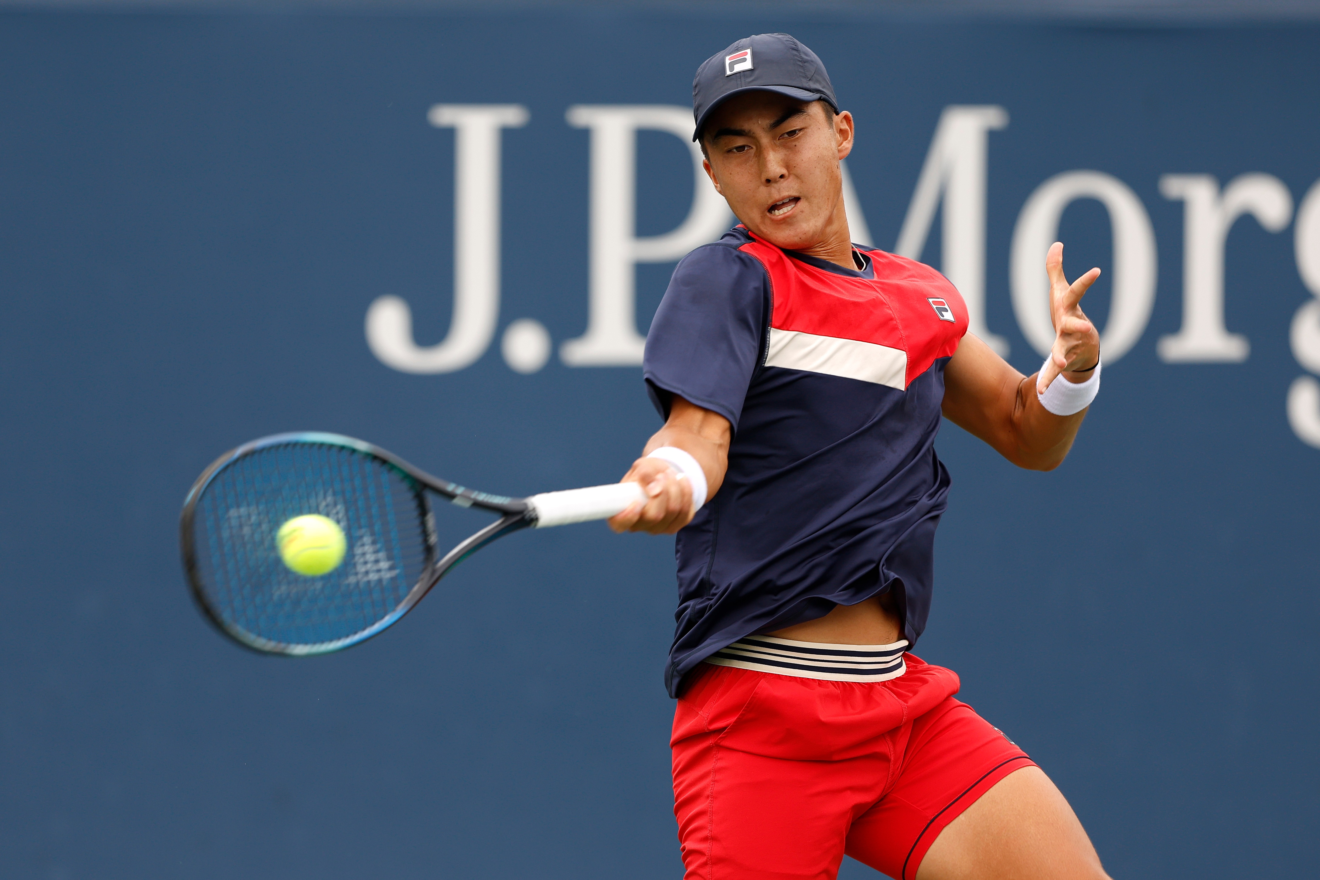 A tennis player wearing a red, white and dark blue shirt and shorts hits a forehand.
