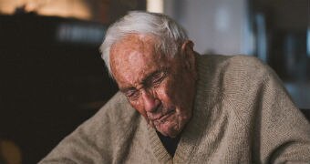 An elderly man in a tanned cardigan sits in a chair with his eyes closed against a dark background.
