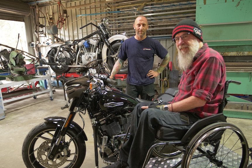 Rob Rees sits in wheelchair next to a black motorcycle, with Brendon Flower standing in the foreground.