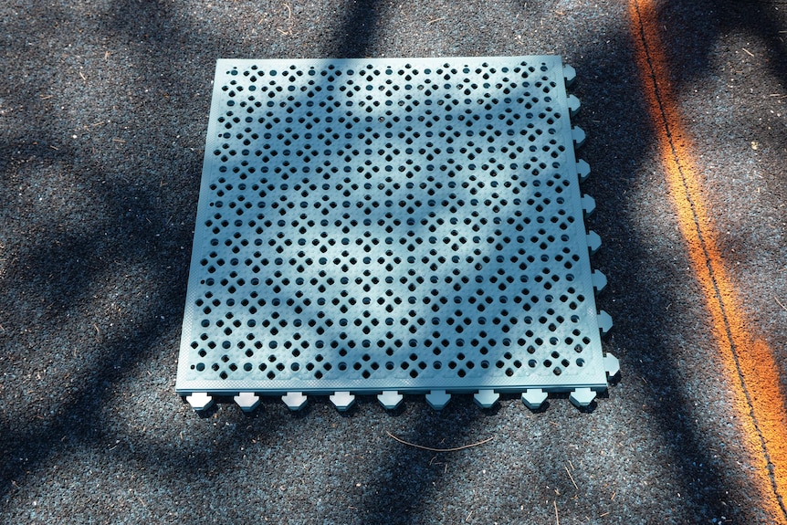 A tile of blue plastic-rubber material sits atop the current playground surface. 
