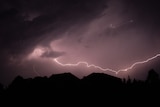 A silhouette picture of houses against a dark purple storm storm cloud with lighting flashing horizontally across the shot.