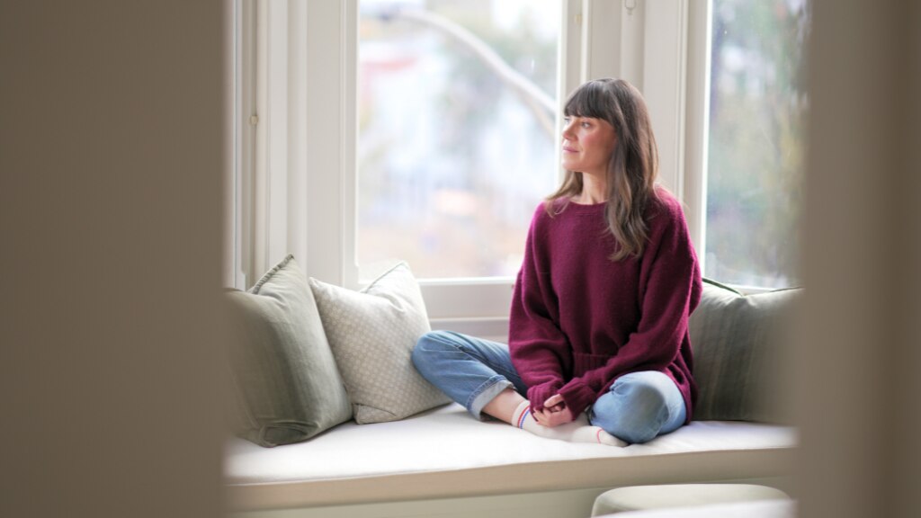 Woman with brown hair and wearing a purple sweater and jeans sits on a window seat looking outward