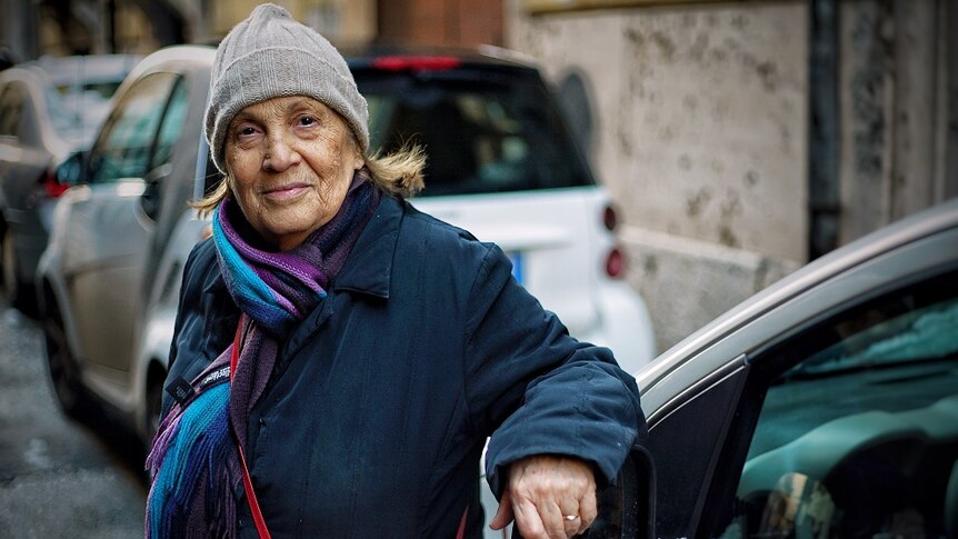 An elderly woman leans against a car's rear view mirror in an Italian street.