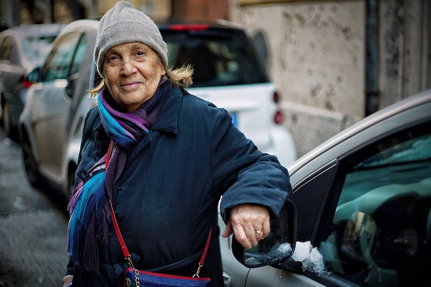 An elderly woman leans against a car's rear view mirror in an Italian street.