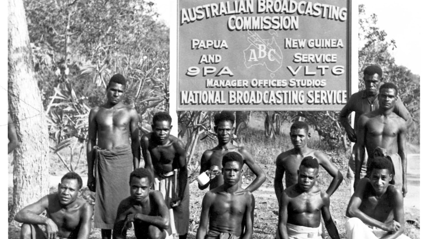 11 shirtless PNG men stand or squat next to sign saying: ABC Papua & New Guinea Service 9PA/VLT6 Manager Offices Studios.