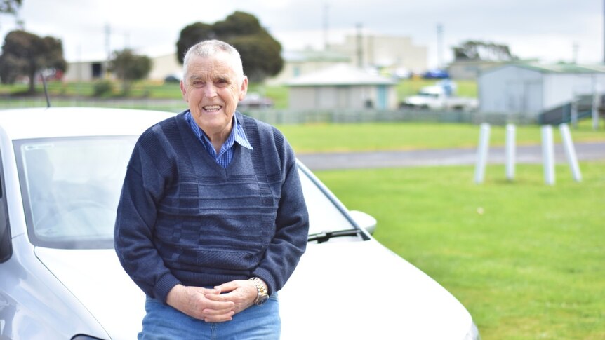 A grey haired man in a blue jumper leans on the bumper of a car