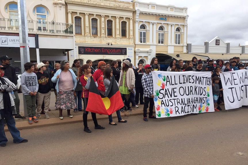 A line of people stands on a road in Kalgoorlie holding banners and an Aboriginal flag.