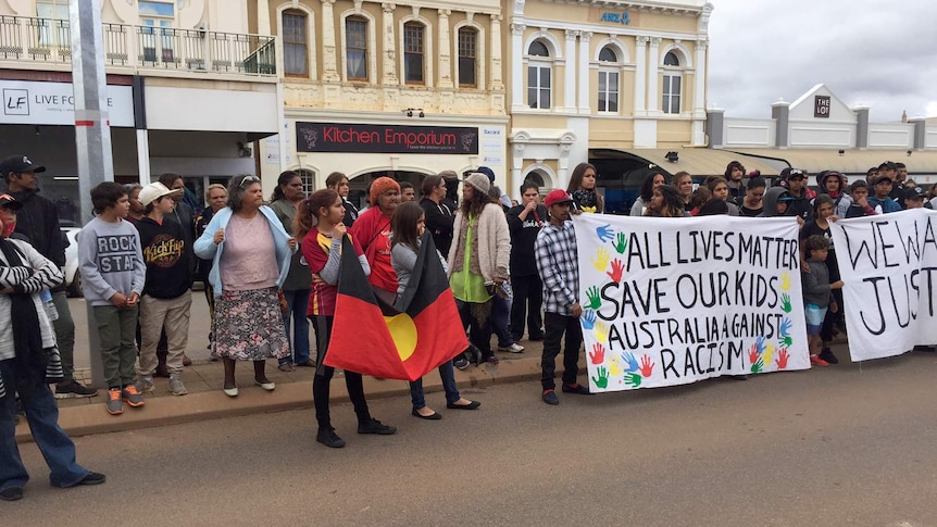 A line of people stands on a road in Kalgoorlie holding banners and an Aboriginal flag.