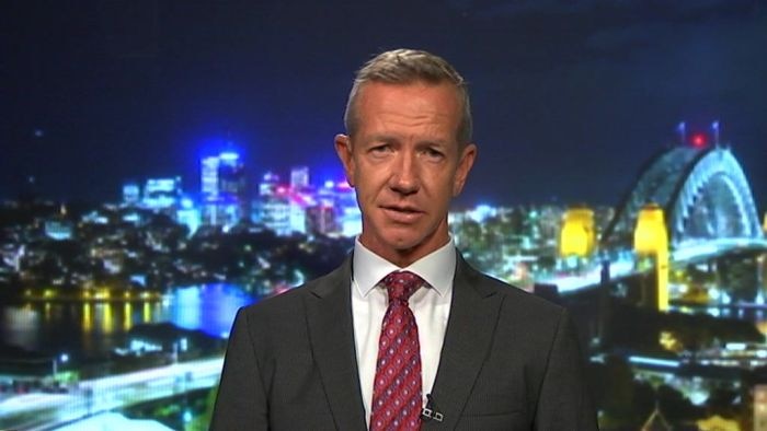 A suited man speaks to the camera. The backdrop of a city at night is behind him.