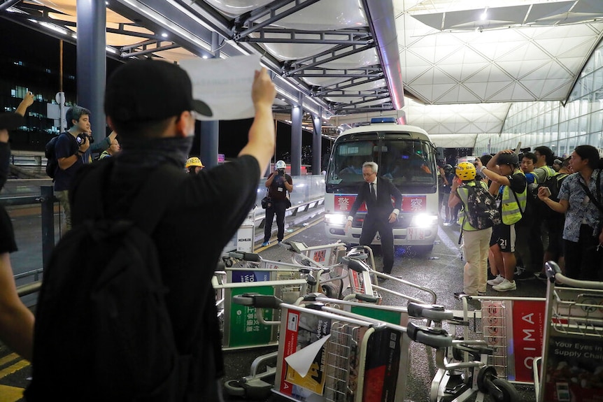 On the road outside Hong Kong Airport, you view a line of overturned trolleys blocking a bus as protesters watch on.
