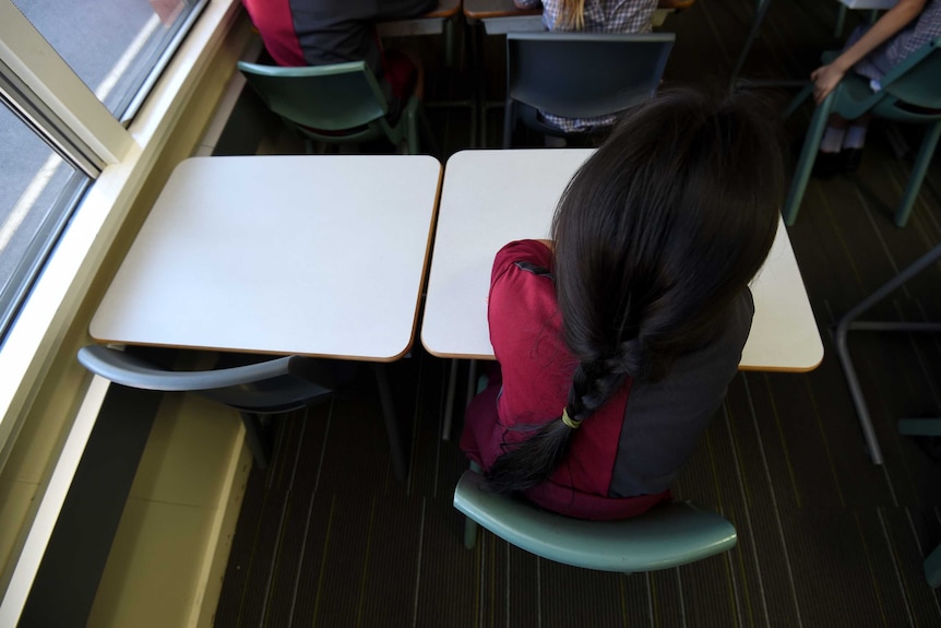 An unidentified child sits alone at a desk in a primary school classroom