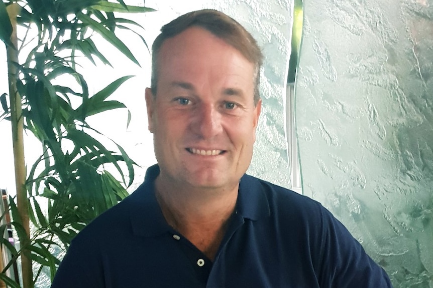 A man with brown hair smiles. He is wearing a navy polo shirt, dark pants and watch.