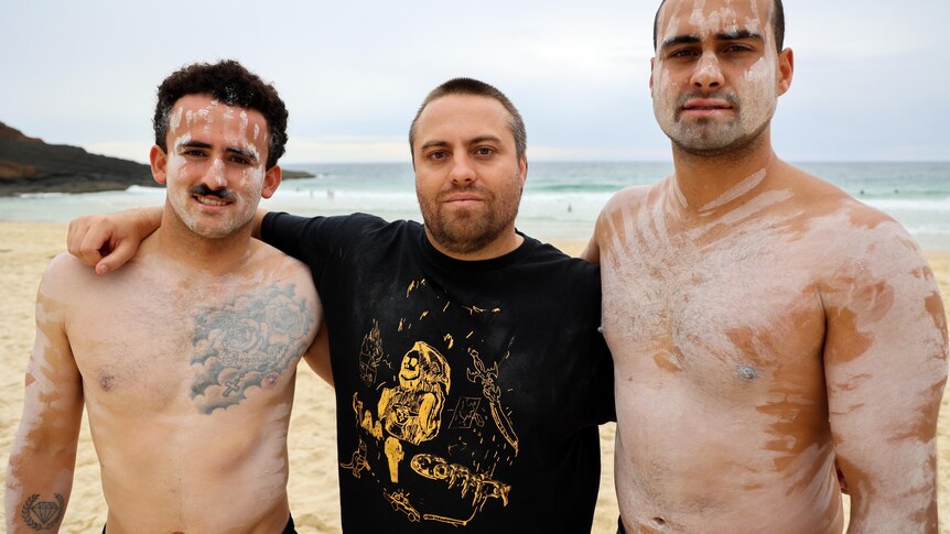 Three surfers stand on the beach looking at the camera