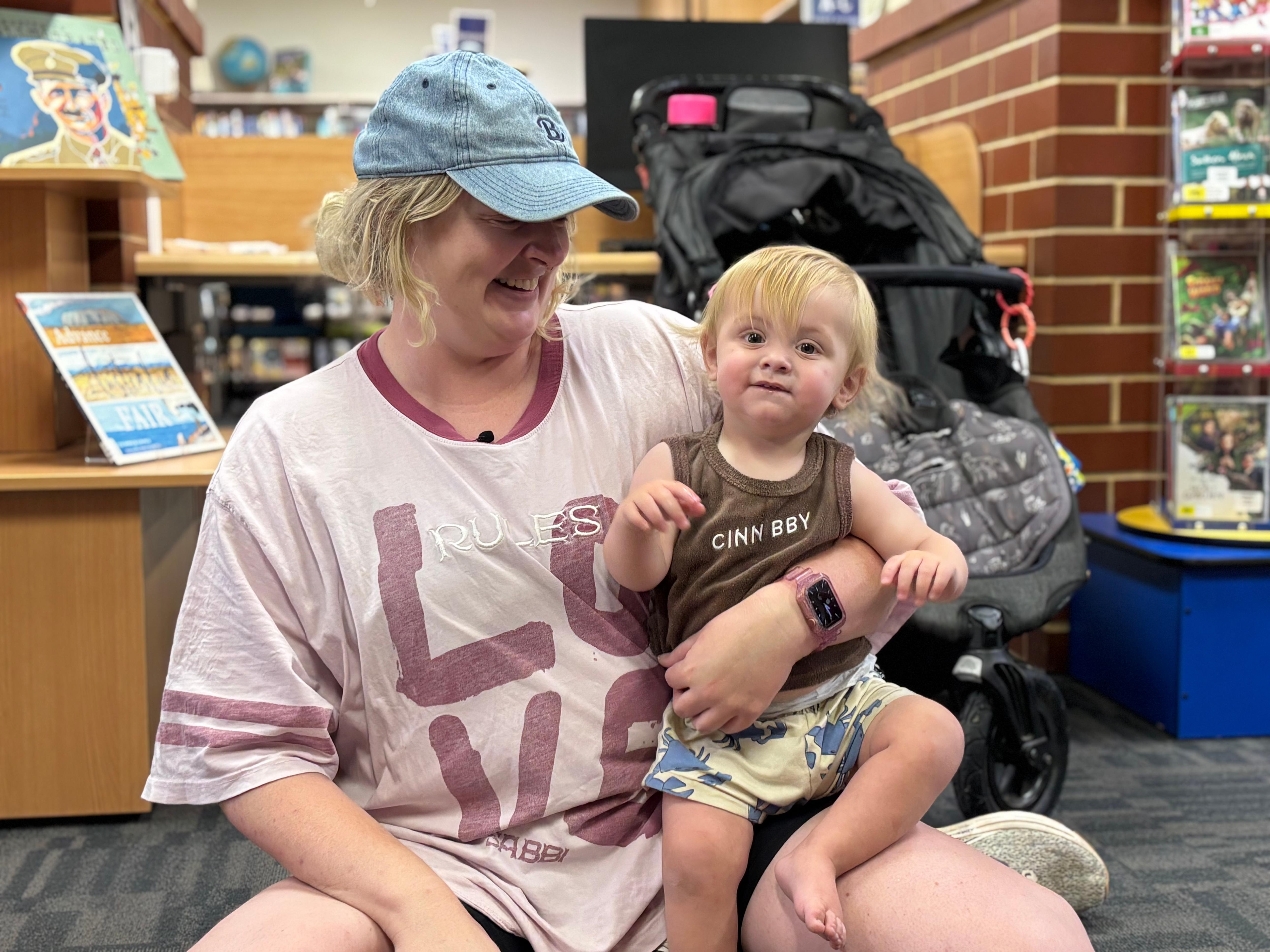 Uma mulher de boné segura uma criança pequena no colo enquanto está sentada em frente a um carrinho de bebê dentro de uma biblioteca.