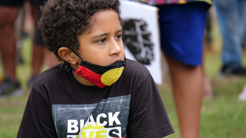 Walali Hatfield, with dark brown curly hair wears an Aboriginal flag face mask, Black Lives Matter shirt with a serious face.