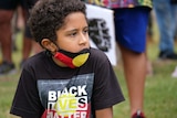 Walali Hatfield, with dark brown curly hair wears an Aboriginal flag face mask, Black Lives Matter shirt with a serious face.