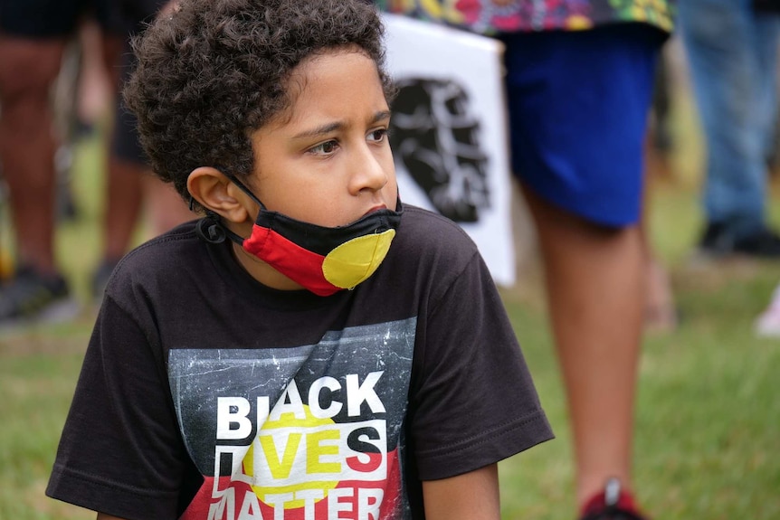 Walali Hatfield, with dark brown curly hair wears an Aboriginal flag face mask, Black Lives Matter shirt with a serious face.