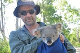 A man in khaki shirt and bucket hat holds a koala wrapped in a blanket with a tagged ear, by a roadside in a forest.