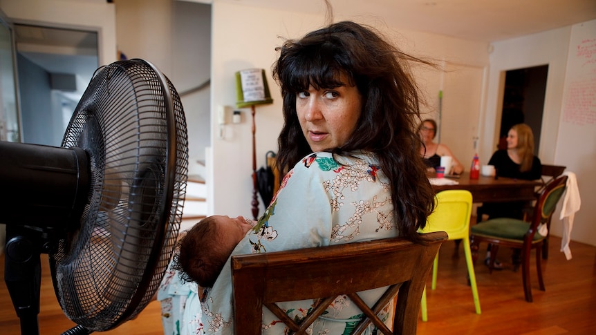 A woman sits on a chair in front of a fan with her baby in her lap, two women sit nearby at a table, for story on share housing.