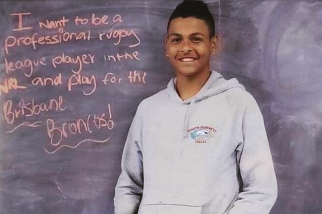 Selwyn Cobbo de niño parado frente a una pizarra con un mensaje que dice que quiere jugar para los Broncos.