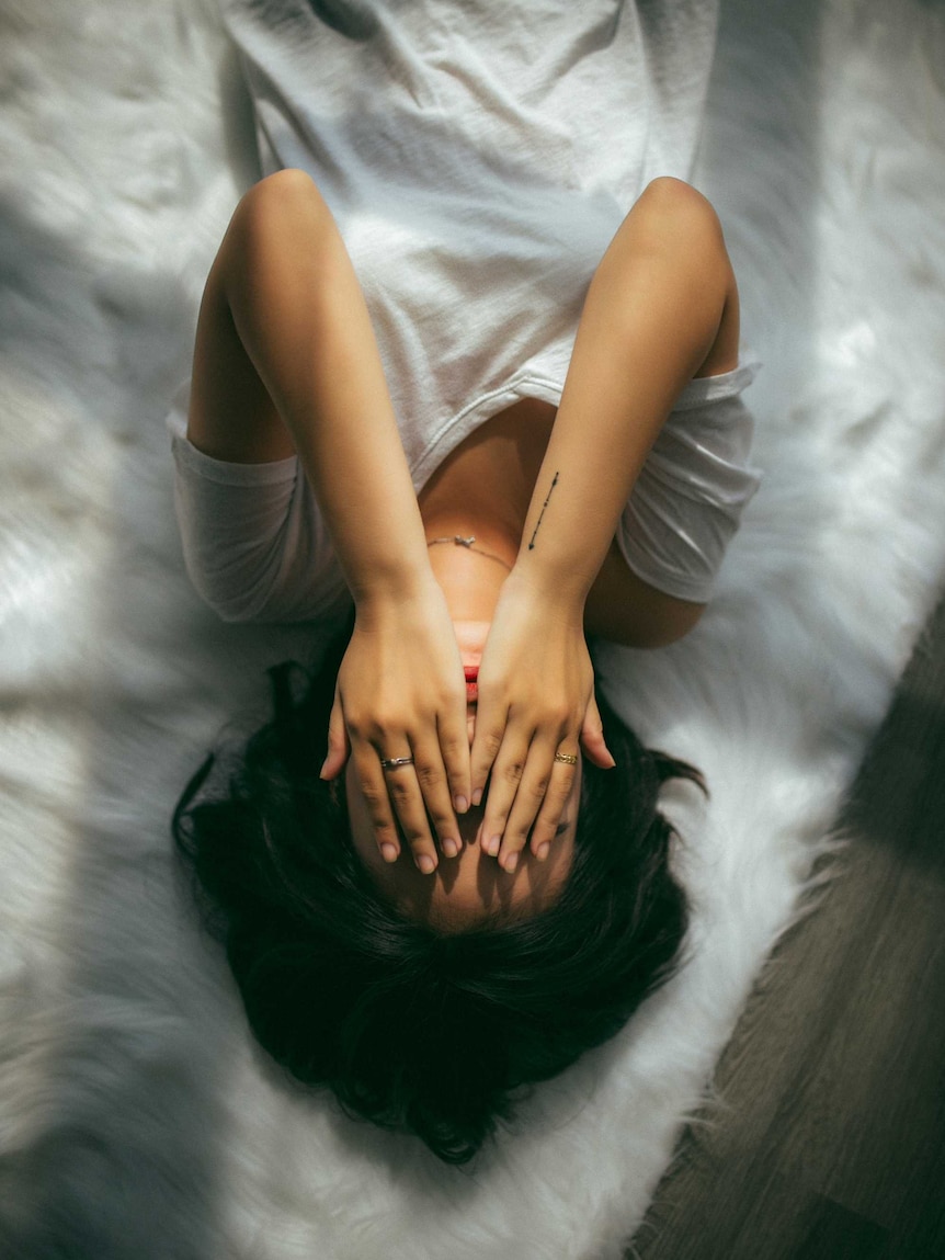 A woman lies on a bed with her hands over her face.