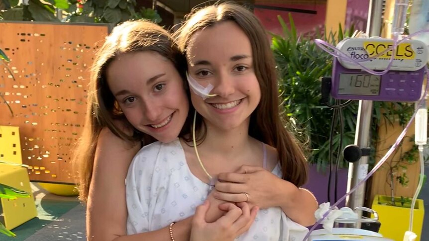 Stella Artuso stands smiling in a hospital courtyard holding an IV drip while her sister Sienna puts her arms around her.