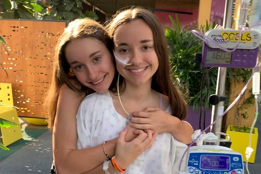 Stella Artuso stands smiling in a hospital courtyard holding an IV drip while her sister Sienna puts her arms around her.