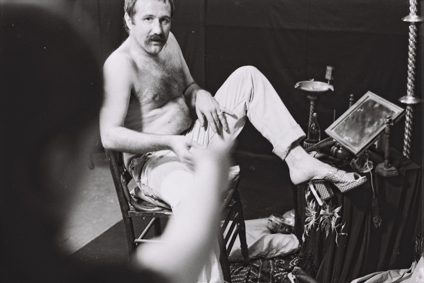 A shirtless man with a moustache sits in a relaxed pose in a cluttered room.