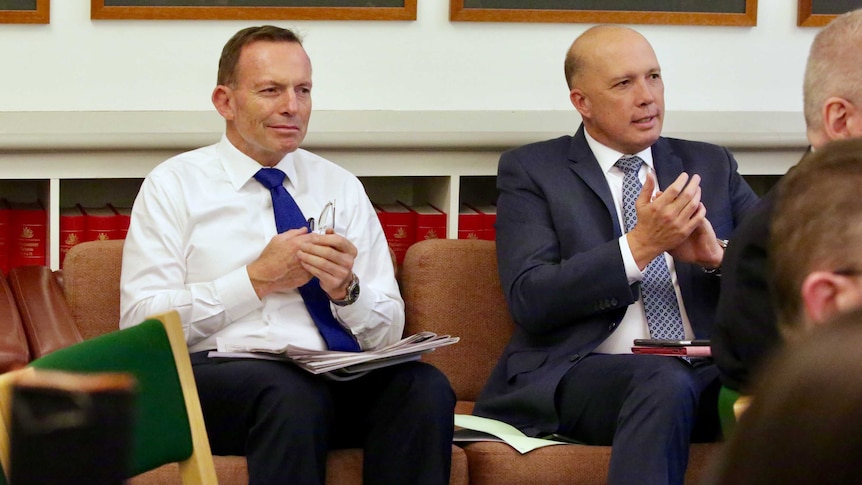 Former PM Tony Abbott, smirking, sits next to Peter Dutton, smiling, in a joint party room meeting