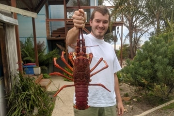 Smiling young man in shorts and t-shirt holding a large dead crayfish towards the camera.