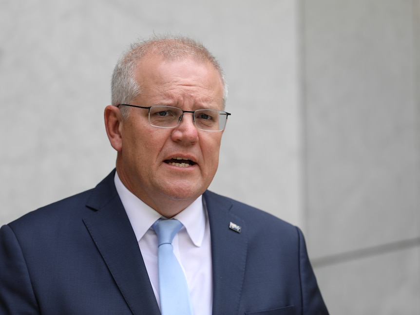 A man with short white hair and glasses wearing a suit and tie speaking at a lecturn