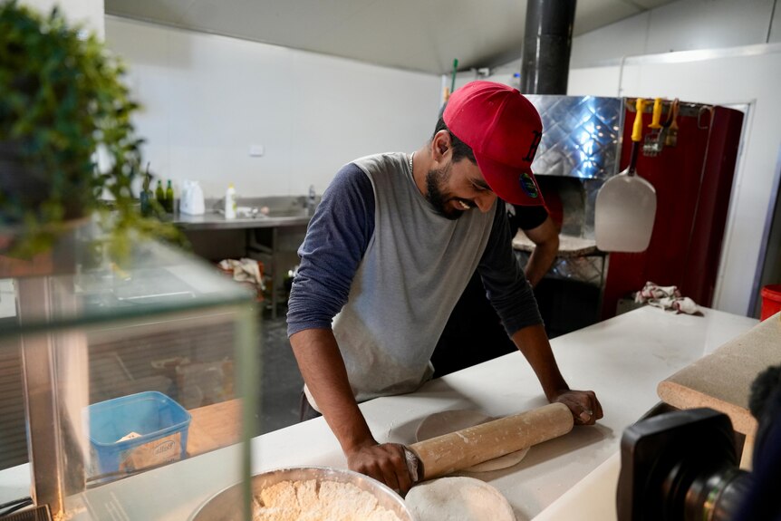 Mudassir Siddique in a grey shirt and red cap rolling pizza dough in a commercial kitchen.