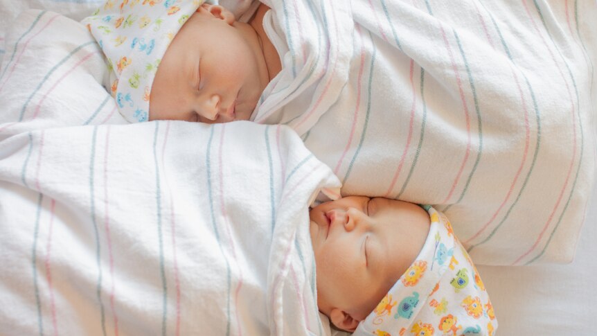 Two twin infants sleeping head to foot, wrapped in blankets and wearing similar hats