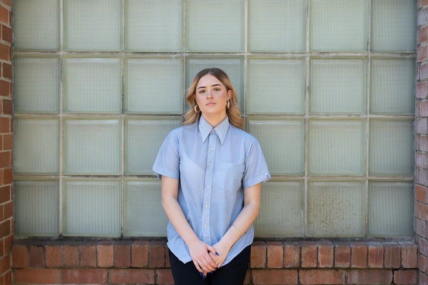 A young woman leans against a glass brick wall her hands together and a neutral expression on her face.