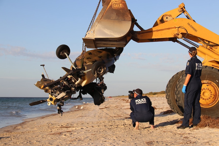 A crane lifts the mangled remains of a gyroscope off a beach, watched by two police officers.