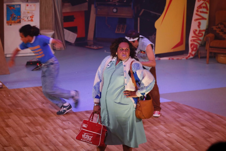 An Indigenous woman stands in the middle of a stage looking stroppy, two boys run in the background