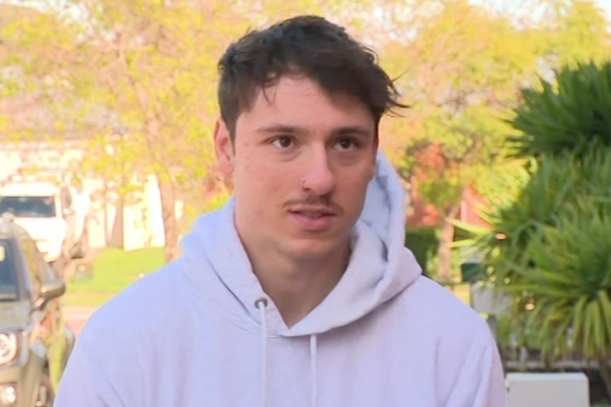 A young man with short dark hair wears a white hooded jumper as he talks to media