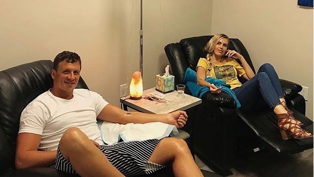 Ryan Lochte sits in a chair with an iv in his arm. Next to him is his wife, also with an iv.