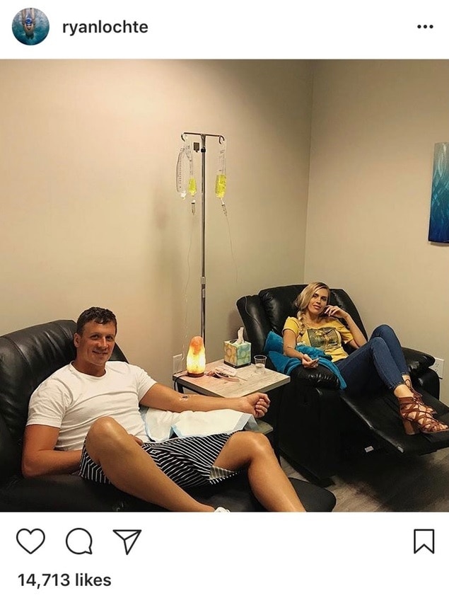 Ryan Lochte sits in a chair with an iv in his arm. Next to him is his wife, also with an iv.