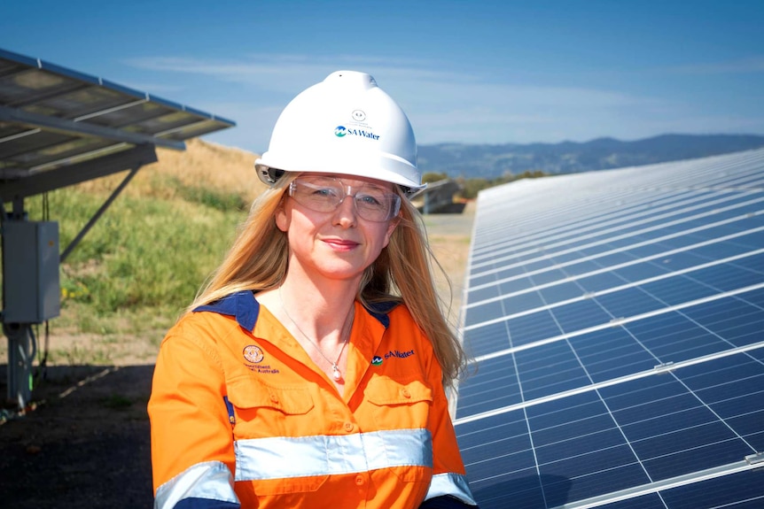 A woman in a high visibility vest and a hard hat stands in front of a solar panel.